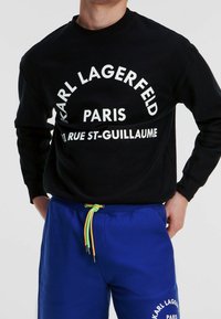 Sudadera negra con estampado blanco que dice "KARL LAGERFELD PARIS" y "11 RUE ST-GUILLAUME", combinada con pantalones deportivos azules con cordón de colores.