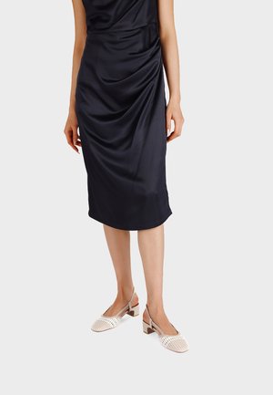 Donna che indossa un vestito midi in raso nero con dettaglio arricciato e scarpe slingback in rete beige con tacco basso su sfondo semplice.