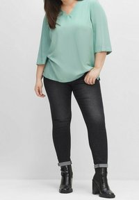Blusa verde menta a maniche corte con scollatura a V, abbinata a jeans scuri aderenti e stivali neri alla caviglia con tacco spesso.