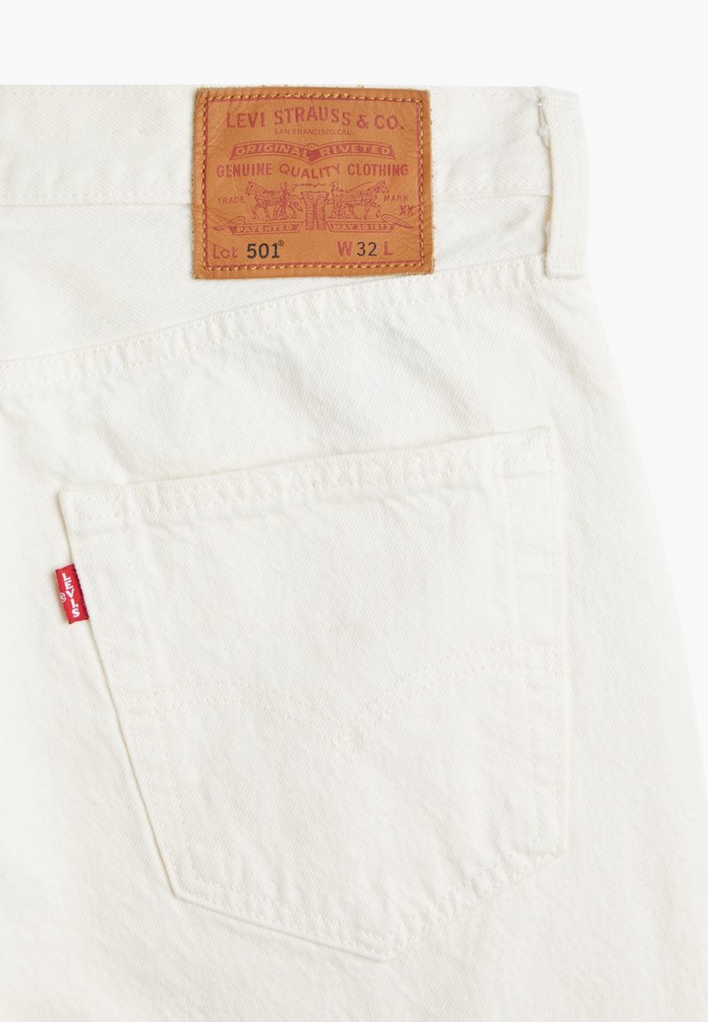 Weiße Denim-Jeans mit einem braunen Lederpatch, das erhabenen Text zeigt. Rotes Levi's-Label an der Gesäßtasche, markante Steppdetails auf dem Stoff.