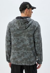 Sudadera con capucha de camuflaje gris de tejido texturizado, con bolsillo frontal, capucha con cordón y puños acanalados, usada con pantalones negros.