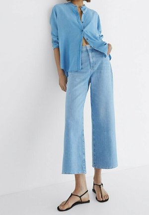 Femme portant une chemise bleu clair boutonnée, un jean bleu clair à jambes larges et des sandales plates noires à fines lanières, debout contre un mur blanc.