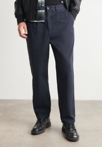 Navyblauwe katoenen broek met een relaxed fit, voorzien van zijzakken, een knoopsluiting en gedragen met zwarte leren schoenen.
