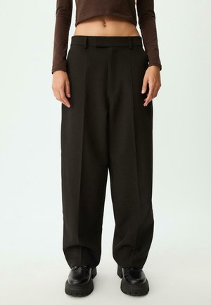 Persoon draagt losse zwarte broek, zwarte dikke schoenen en een cropped bruine top met lange mouwen tegen een effen achtergrond.