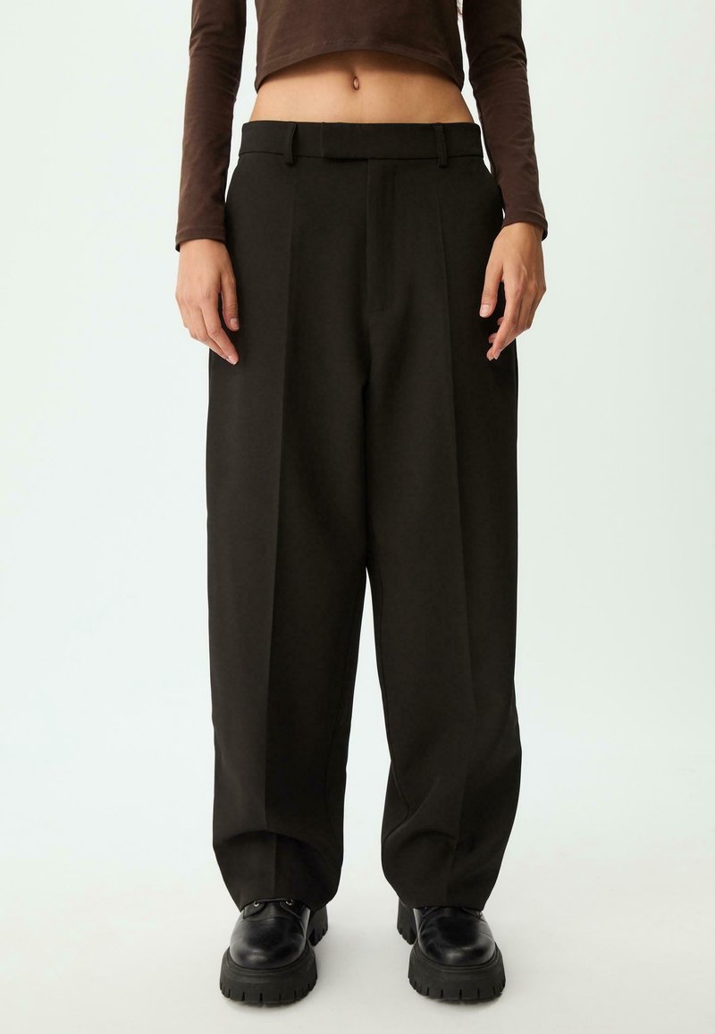 Personne portant un pantalon noir ample, des chaussures noires épaisses et un haut marron à manches longues court sur un fond uni.