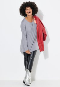 Graues Langarmoberteil über schwarzen Lederleggings. Das Modell hält eine rote Lederjacke. Silberne Cowboystiefel mit strukturierten Details, vor einem weißen Hintergrund.