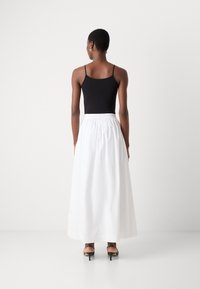 Bodysuit sans manches noir associé à une jupe blanche longue jusqu'au sol avec une taille élastique. Modèle portant des talons noirs, vu de derrière.