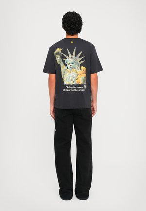 Camiseta de algodón negra con un gráfico de un gato junto a la Estatua de la Libertad, rodeado de ositos de peluche, con texto debajo.