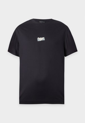 Bărbat purtând tricou negru cu logo mic, blugi largi albastru închis și pantofi sport albi, stând cu o mână în buzunar.