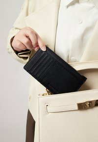 Portafoglio nero con texture, dotato di slot per carte e cerniera, parzialmente inserito in una borsa color crema con dettaglio della chiusura dorata.