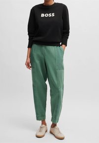 Černá mikina s nápisem "BOSS" v bílé barvě, kombinovaná se zelenými kalhotami ve stylu utility s bočními kapsami a zúženými manžetami. Béžové tenisky.