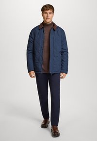 Chaqueta de navy acolchada con cuello marrón, combinada con un suéter de cuello alto marrón y pantalones navy. Presenta botones y una textura suave. Botas de cuero marrón.