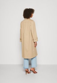 Un trench coat bej cu o croială lejeră, cu mansete cu nasturi și un detaliu pliant la spate, asortat cu blugi largi de culoare albastru deschis și sandale cu toc de culoare marron.
