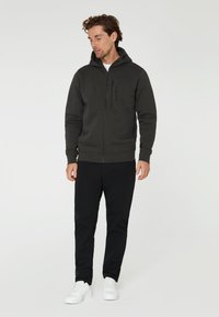 Dunkelgrauer Reißverschluss-Hoodie mit Kapuze, der eine glatte Textur, einen Frontreißverschluss und Logodetails aufweist. Kombiniert mit schwarzen Hosen und weißen Sneakers.