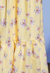 Žltá tkanina s fialovými, oranžovými a bielymi kvetinovými vzormi, ktorá má zbernú ševovú funkciu na svetlo modrom pozadí.