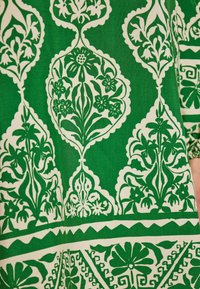 Țesătură verde cu modele florale albe intricate, prezentând motive stilizate și margini geometrice, creând un efect vizual texturat și stratificat.
