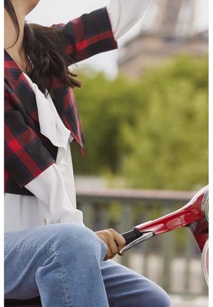 Persona con chaqueta de cuadros y camisa blanca sentada en un scooter rojo, sosteniendo el manillar, con un fondo de vegetación borrosa.