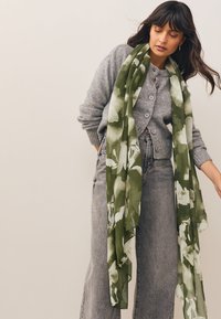 Sciarpa floreale verde con texture a coste, drappeggiata su un cardigan grigio. Tessuto morbido, design leggero e motivo floreale intricato.