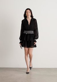 Black tiered dress with long sleeves, pleated texture, silver-striped waistband, and ruffled hem. Paired with black pointed-toe shoes.
