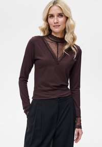 Brązowa bluzka z długim rękawem, ozdobiona koronkowym kołnierzykiem i koronkowymi detalami na rękawach. Gładka tkanina o dopasowanym kroju. W zestawieniu z czarnymi spodniami.