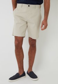 Pantalones cortos de algodón beige con corte recto, bolsillos delanteros y un cierre de un solo botón. Combinados con zapatos navy sin cordones.