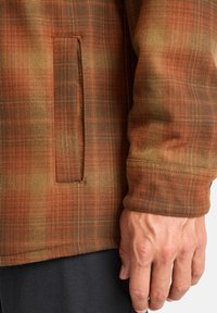 Chaqueta de cuadros en tonos naranjas y marrones, con un bolsillo lateral y una tela texturizada. Las mangas ajustadas completan el diseño.