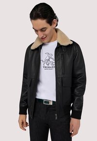 Czarna skórzana kurtka bomber z beżowym futrzanym kołnierzem, zamkiem błyskawicznym z przodu, ściągaczami na mankietach i dwoma dużymi przednimi kieszeniami. Noszona na białej koszulce z nadrukiem.