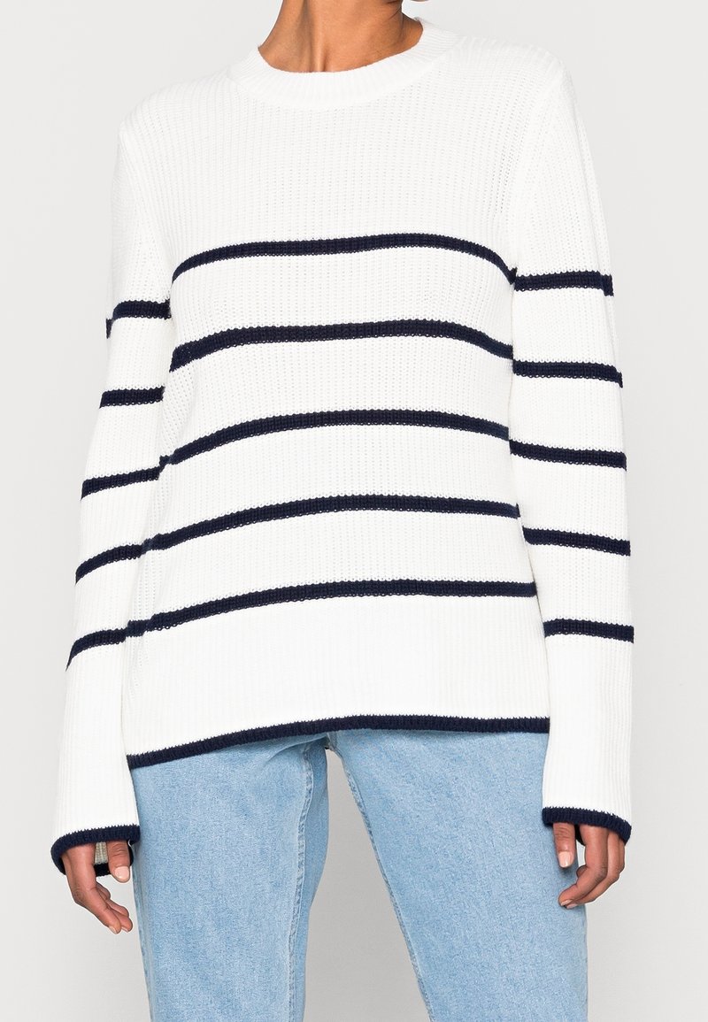 Maglione bianco a maglia con strisce orizzontali blu navy, texture a costine, collo rotondo e maniche lunghe, abbinato a jeans azzurri chiari.