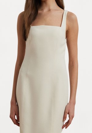 Femme portant une robe ajustée sans manches, à encolure carrée, de couleur blanc cassé, sur un fond clair uni.