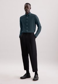 Maglietta a maniche lunghe color teal con colletto classico e bottoni bianchi, abbinata a pantaloni tapered neri e scarpe stringate nere. Texture liscia.