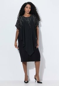 Femme aux longs cheveux bouclés portant une robe noire jusqu'aux genoux avec un voile transparent en perles et des escarpins noirs à talons pointus avec des embellissements décoratifs.