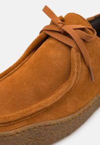 Scarpa in suede marrone con suola texturizzata, caratterizzata da una forma arrotondata, design con lacci e parte superiore liscia con dettagli di cucitura minimi.