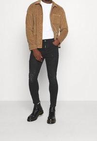 Hombre llevando una chaqueta de pana color beige, camisa blanca, jeans negros ajustados rasgados y botas de cuero negras, de pie con una mano en el bolsillo.