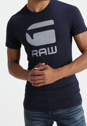 Mand iført en mørkeblå T-shirt med et stort gråt logo og ordet "RAW" over brystet, hænderne samlet foran.