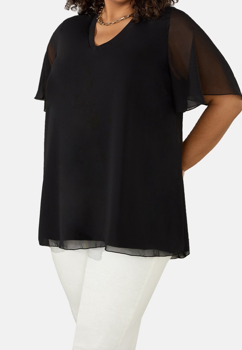 Svart chiffongblus med V-ringning och genomskinliga volangärmar, med en lös passform och en något längre bakstycke, kombinerad med vita byxor.