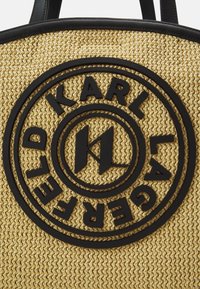 Sac en toile beige tissé avec logo en caoutchouc noir présentant un design circulaire et du texte. Matériau résistant, doubles poignées et surface texturée.