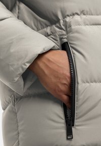 Lichtgrijze pufferjack met een ritsvak. De stof heeft een gladde textuur, en de rits is zwart met een opvallende trekker.