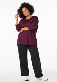 Weinrotes Langarmshirt mit Rundhalsausschnitt, kombiniert mit schwarzen weiten Hosen. Das Outfit wird durch weiße Sportschuhe ergänzt.