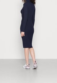 Femme portant une robe midi à manches longues côtelée bleu marine et des baskets Nike Air blanches et roses, debout devant un fond blanc uni.