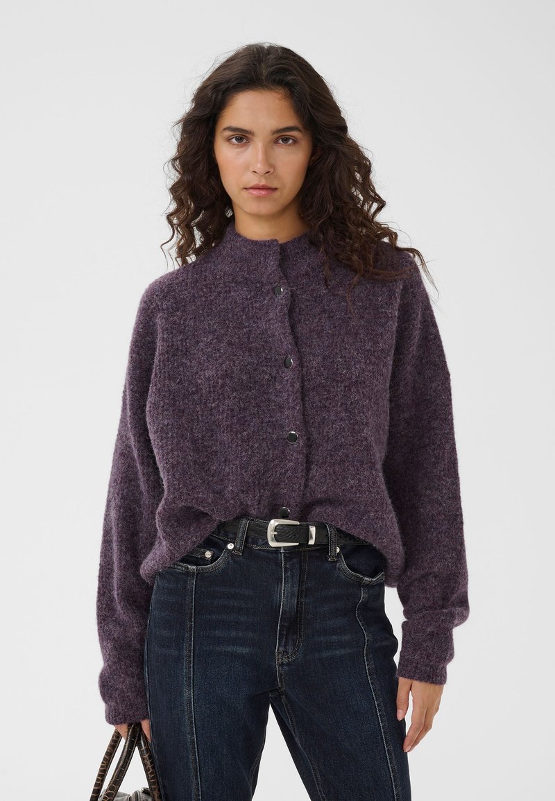 Femme aux cheveux bouclés portant un pull en laine violet boutonné, un jean foncé avec une ceinture noire, tenant un sac à main d'une main.