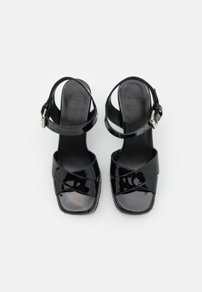 Sandalias de tacón de charol negro con correas cruzadas, punta cuadrada, cierre de hebilla en el tobillo y textura suave. Presenta un tacón ancho.