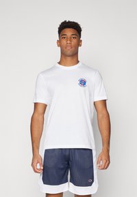 T-shirt en coton blanc avec col rond et manches courtes, présentant un motif de basket-ball bleu sur la poitrine. Porté avec un short en maille bleu marine.