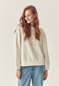 Jasno beżowy sweatshirt z kapturem, z ribbowanymi mankietami i luźnym krojem. Akcentowany sznurkiem i minimalistycznym designem. W zestawie z niebieskimi dżinsami.