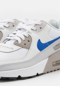 Nike Air Max tenisice u bijeloj i sivoj koži s plavim naglaskom. Imaju teksturirane panele, dizajn s vezicama i vidljivu zračnu amortizaciju u potplatu.