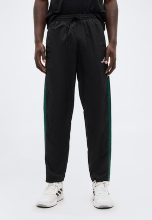 Personne portant un pantalon de survêtement noir avec des bandes latérales vertes et des baskets blanches à rayures noires, debout devant un fond blanc uni.