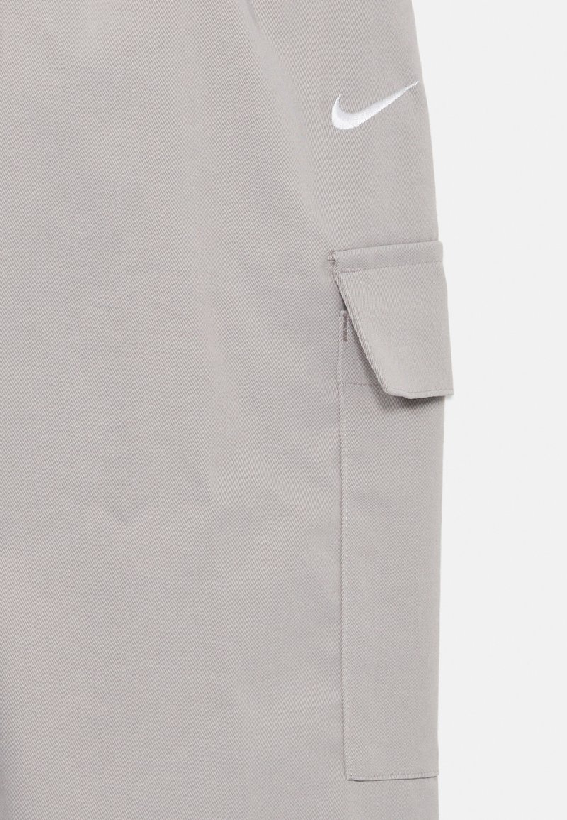 Helle graue Nike Cargo-Hosen besitzen eine Seitentasche mit Klappe und bestehen aus weichem, strukturiertem Material. Das Nike-Logo ist in Weiß dargestellt.