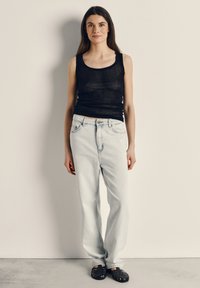 Mladá žena stojaca, oblečená v čiernom tielku bez rukávov, svetlomodrých džínsach a čiernych pletených topánkach, na pozadí s jednoduchou stenou.