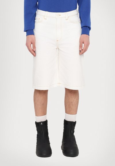 Pantalones cortos de mezclilla blanca con corte recto, con costuras amarillas, bolsillos frontales y cinturilla. Combinados con botas negras con cremallera y calcetines blancos.