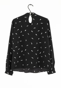 Blouse noire à manches longues avec un motif de plumes blanches, fermeture à l'arrière en forme de clef, et poignets boutonnés. Tissu doux avec une coupe décontractée.