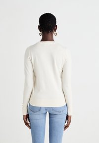 Pull crème à manches longues avec un col rond, coupe ajustée, ourlet côtelé et accents de boutons sur les manches, associé à un jean bleu clair.
